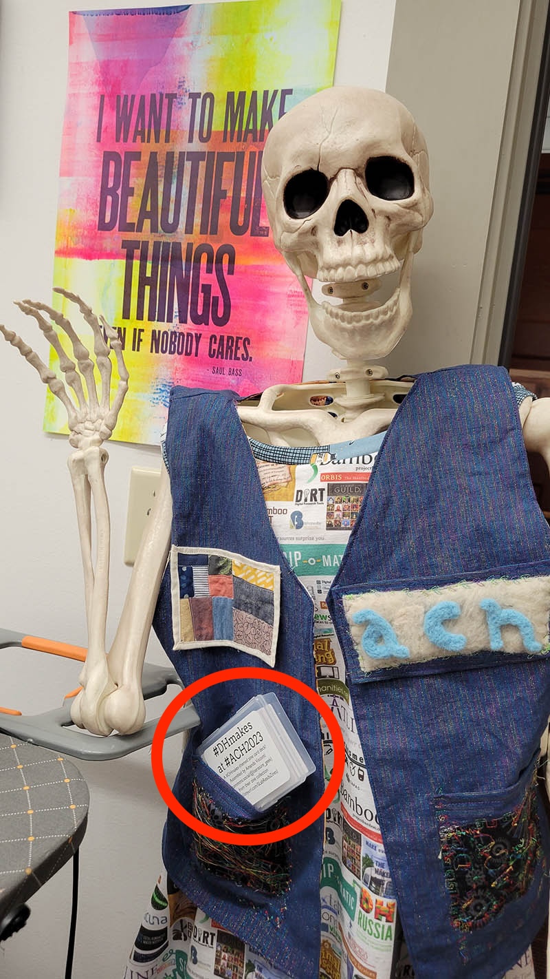 Photo of a fake, adult-size skeleton (Dr. Cheese Bones) wearing the ACH 2023 #DHMakes crew's collaborative DH making vest, which boasts a variety of neat small making projects such as a data visualization quilt patch and felted conference name letters. One of my themed reading card decks is visible half-tucked into its vest pocket. Photo and Dr. Bones appearance by Quinn Dombrowski.