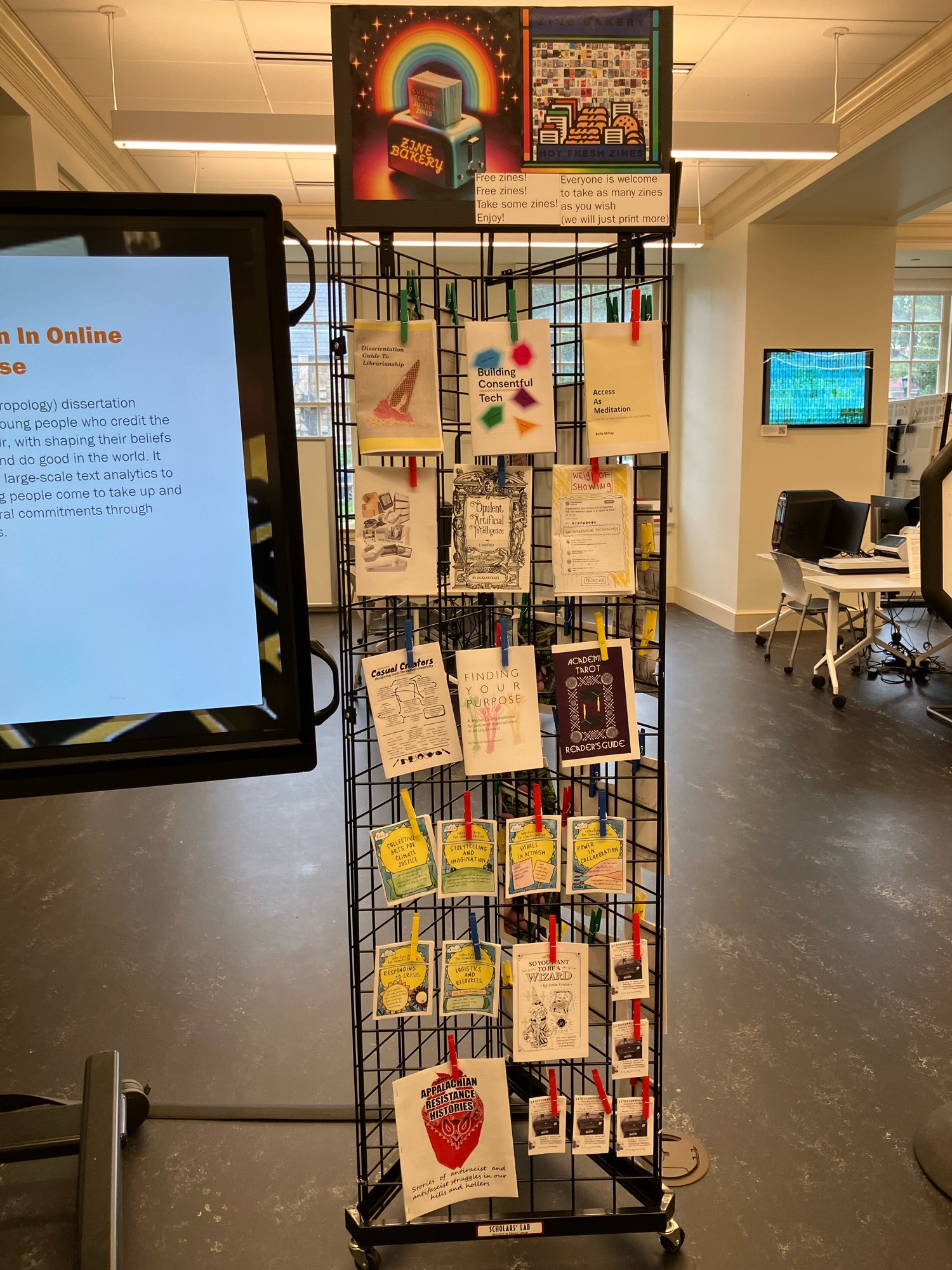 Photo of the Scholars' Lab public space Zine Bakery free zine rack, a tall black metal grid with three sides, with various paper zines held over its surface using variously colored clothespins