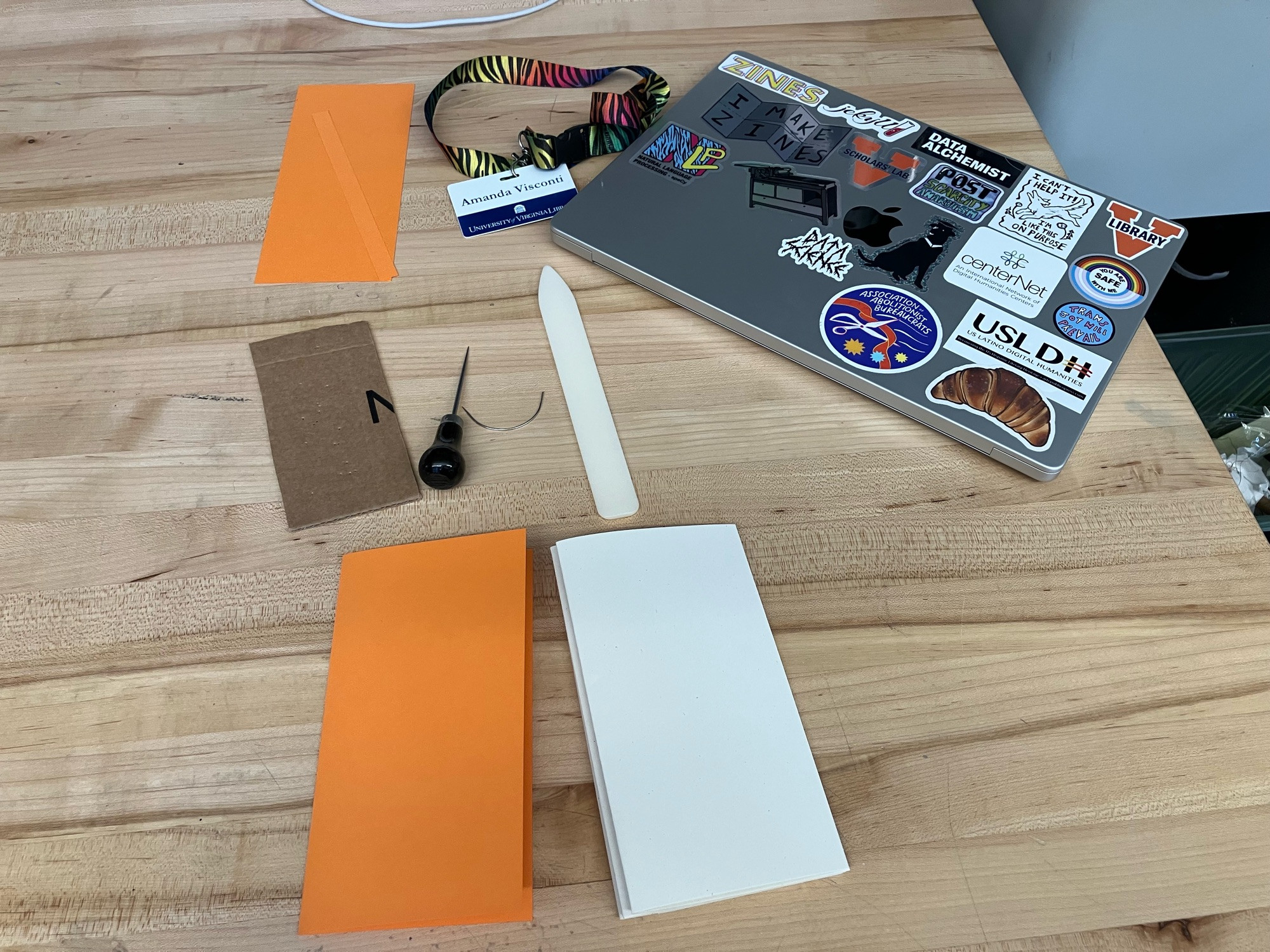 A work table spread with supplies for handmaking a journal, including folded inner paper, an orange construction paper cover, a half-circle needle, cardboard for stabbing the awl into, and a white bone folder to help crease pages at the spine