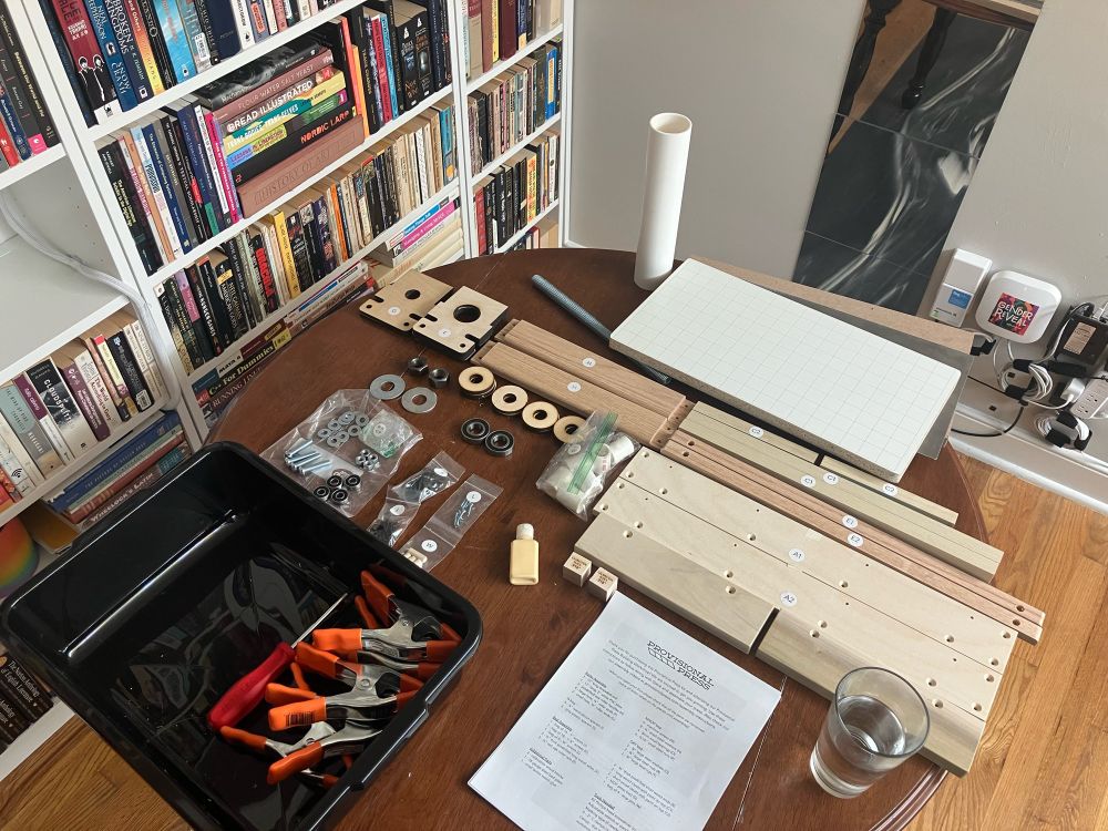 Photo of a kitchen table covered with neatly arranged pieces of a to-be-assembled tabletop letterpress roller printing press. The pieces are mostly wood slats, plus a large PVC-pipe tube, clamps, glue, small metal hardware, and a long thick metal bolt