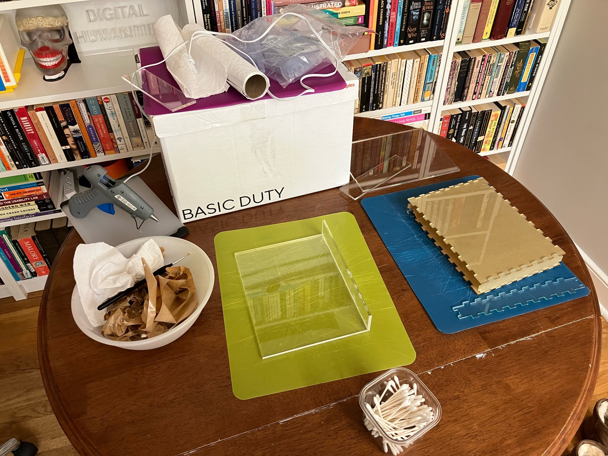 Photo of a kitchen table covered with supplies for gluing acrylic and glass boxes together. Supplies include a hot glue gun, cutting boards, a clear thick acrylic material made into a shape intended to eventually be a box with two sides glued together; a book-shaped rectangular box made of glass, missing the top and bottom sides, where the edges are cut into jigsaw crenellations, with glue drying between the joints; a cup of Q-tips; a bowl of trash; a cardboard box.